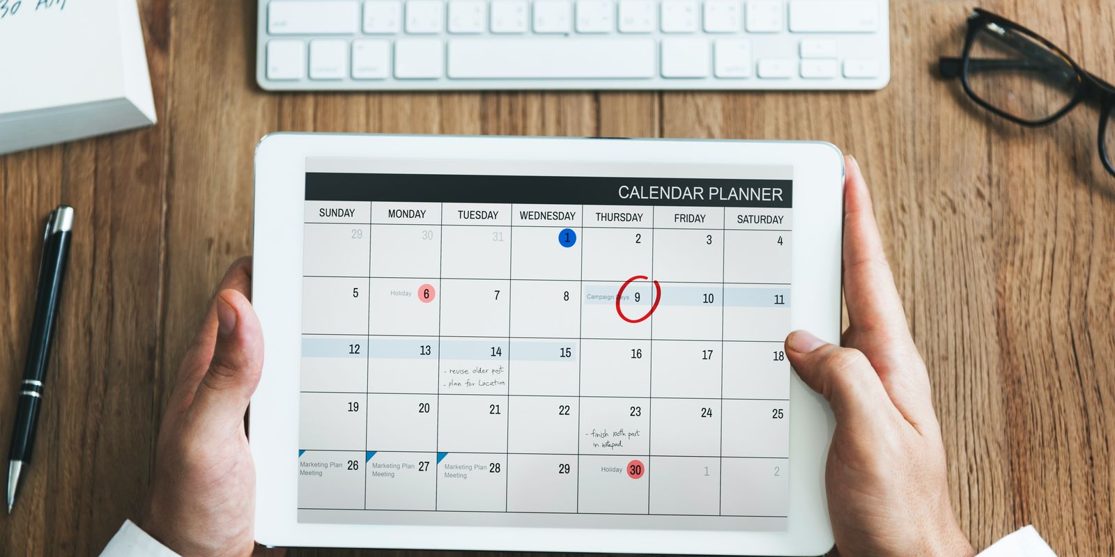 Hands holding a tablet that shows a monthly calendar planner on a wooden desk